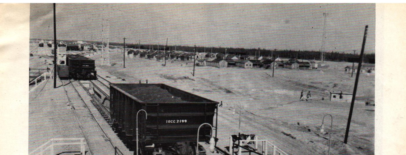 An ore car marked IOCC 2199 on track at the Sept-Îles dock, with rows of catering camp barracks visible on bare ground in the background, July 1954